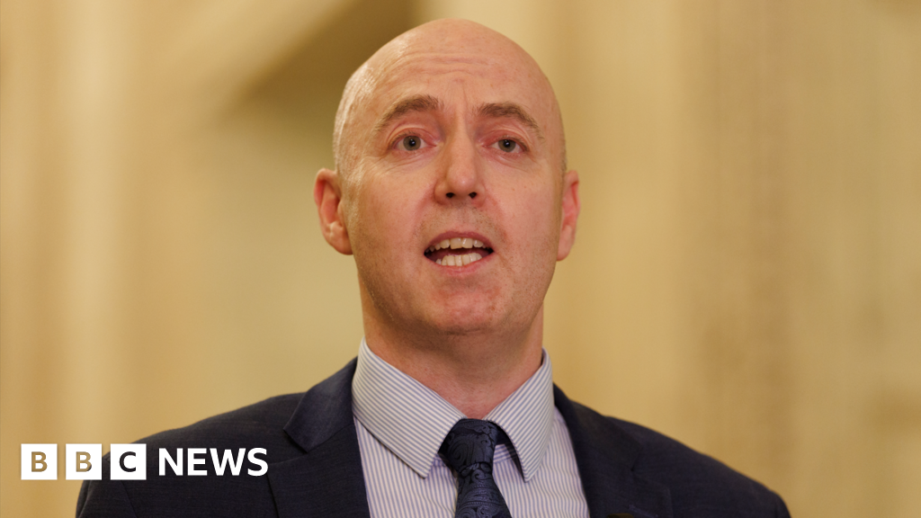 Jon Burrows is talking. He's wearing a navy suit with a white and blue stripped shirt and a navy tie with a pattern. The background is out of focus.
