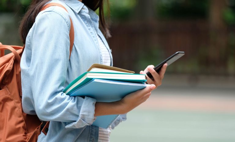 Student holding books and smartphone while walking on campus.