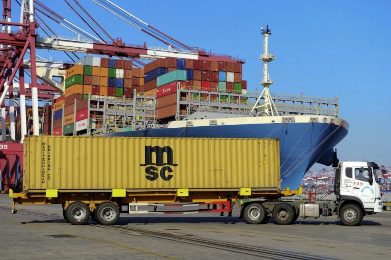 A truck transports a shipping container drives past a loaded cargo ship.