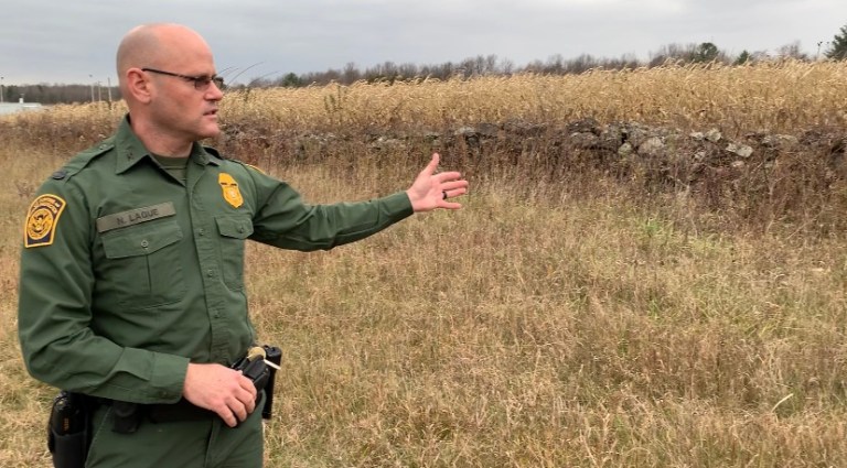 A U.S. Border Patrol agent points to a stone wall, which serves as the boundary between New York and Canada.