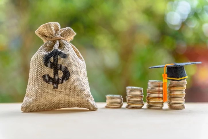 Education expense or student loan for post secondary education concept: Dollar bag, graduation cap on row of coins on a table, depicts loan or money designed to help students pay for associated fees.