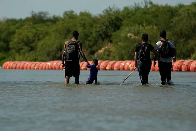 Migrants crossing into the U.S. from Mexico walk along large buoys being used as a floating border barrier on the Rio Grande Tuesday, Aug. 1, 2023, in Eagle Pass, Texas. (AP Photo/Eric Gay)