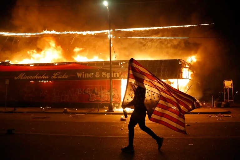 A person runs while holding up a large American flag as a fire burns in the background.