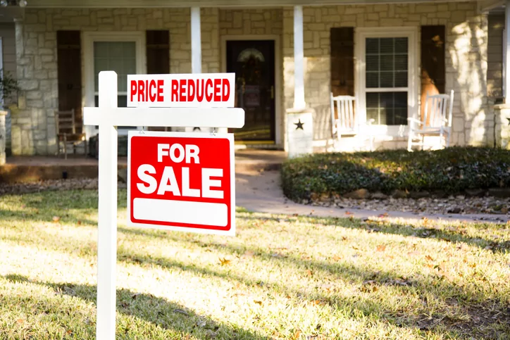 Home for sale with red and white real estate sign during the fall season. Fall season with leaves on ground. Front porch and windows in background. Residential neighborhood. Moving house, relocation concept. Price Reduced.