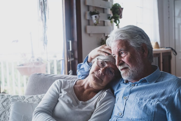 Couple of two sad and depressed old people sitting on the sofa at home looking outside the window. Lockdown and quarantine lifestyle indoor.