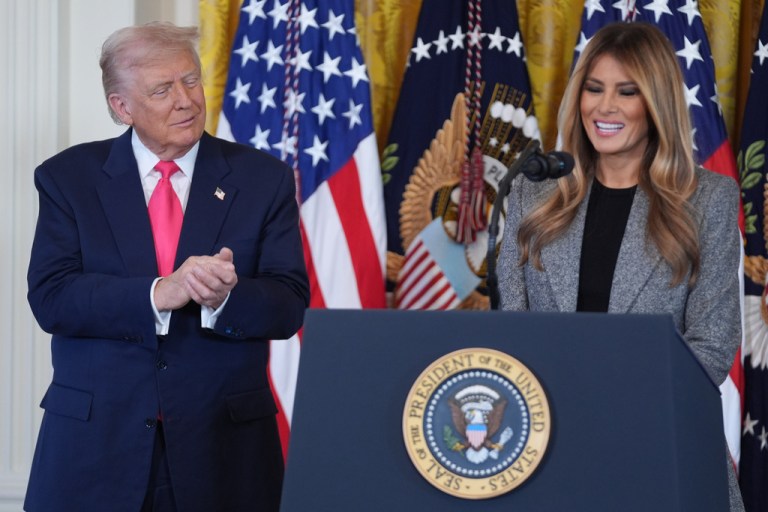 President Donald Trump claps as first lady Melania Trump speaks at a podium.
