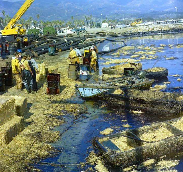 Workmen using pitchforks, rakes, and shovels attempt to clean up oil-soaked straw from a beach.