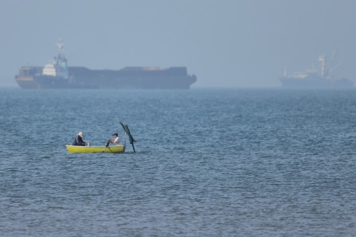 Two men sit in a small boat on the water as cargo ships are anchored in the background in the Strait of Hormuz off Bandar Abbas, Iran, Monday, May 4, 2026.