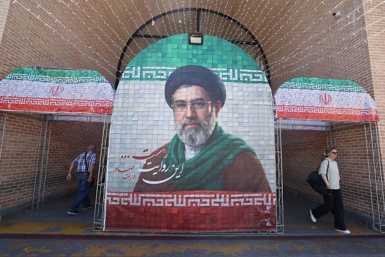 Iranian commuters walks past a large tiled image of Iran's current leader Ayatollah Mojtaba Khamenei.