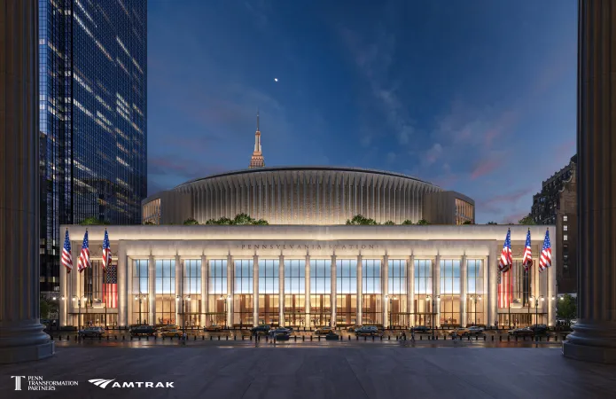 A conceptual nighttime rendering of the proposed redesigned Penn Station viewed from Eighth Avenue, featuring a monumental stone façade, expanded glass entrances, and a reimagined circular arena structure above the station. The image also shows large American flags flanking the entrance and a decorative eagle-style seal visible inside the glass façade, part of the broader architectural vision unveiled by Amtrak and Penn Transformation Partners for the Manhattan transit hub redevelopment. (Rendering courtesy of Penn Transformation Partners and Amtrak; designed by PAU.)